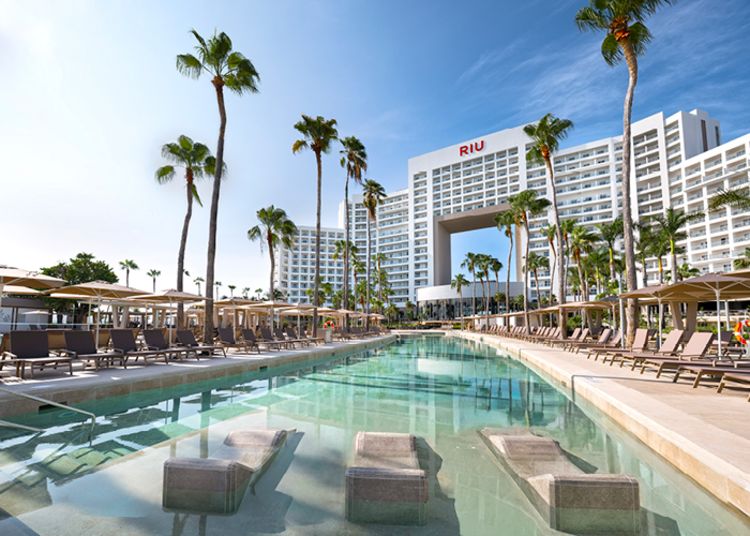 A large swimming pool surrounded by lounge chairs and umbrellas, with a building in the background and a clear blue sky above.