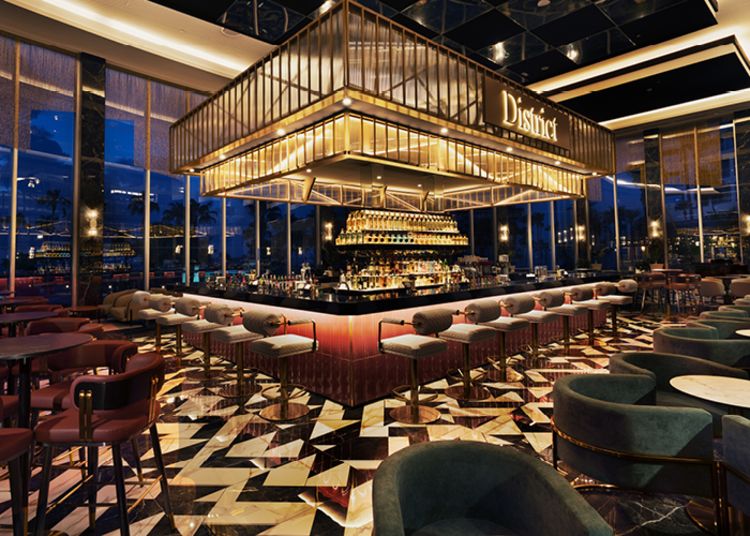 A large room filled with lots of tables and chairs, a bar with bottles on the platform, a name board with text, lights on the ceiling, and glass walls that offer a view of the buildings, trees, and sky outside.