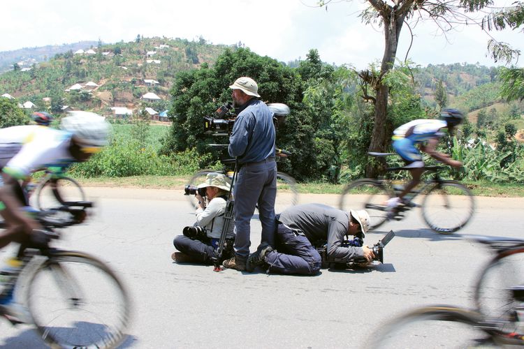 “The Royal Tour” camera crew takes position in the middle of a road to shoot the Rwanda national cycling team zooming past.