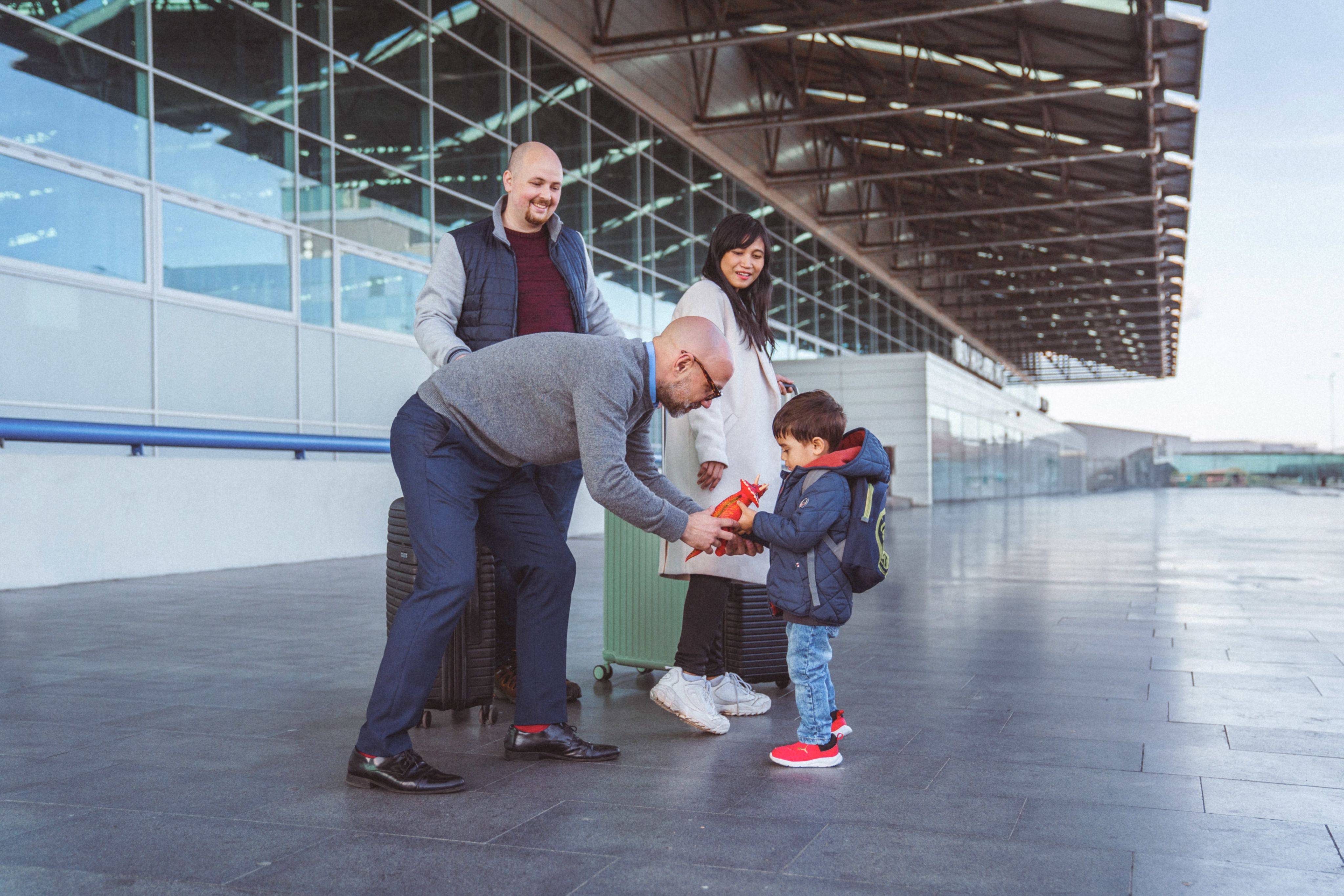 A family of four - a man, a woman, and two children - standing in front of a building with glass walls. The man is holding a trolley bag and the woman is holding an object. The sky is visible in the background.