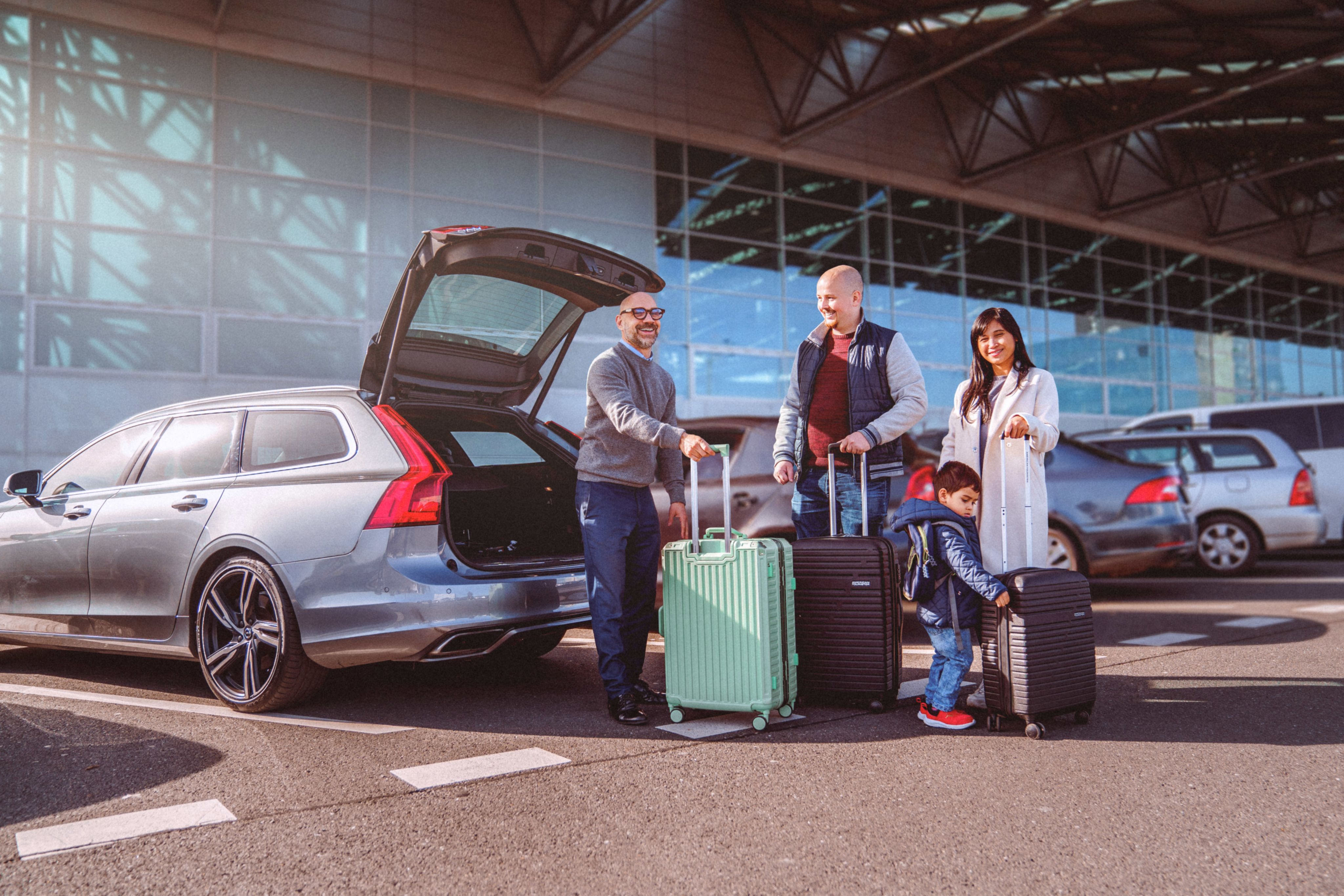 A family of four - a man, a woman, and two children - standing next to a car with their luggage. The car is parked on the side of the road, with a building in the background.