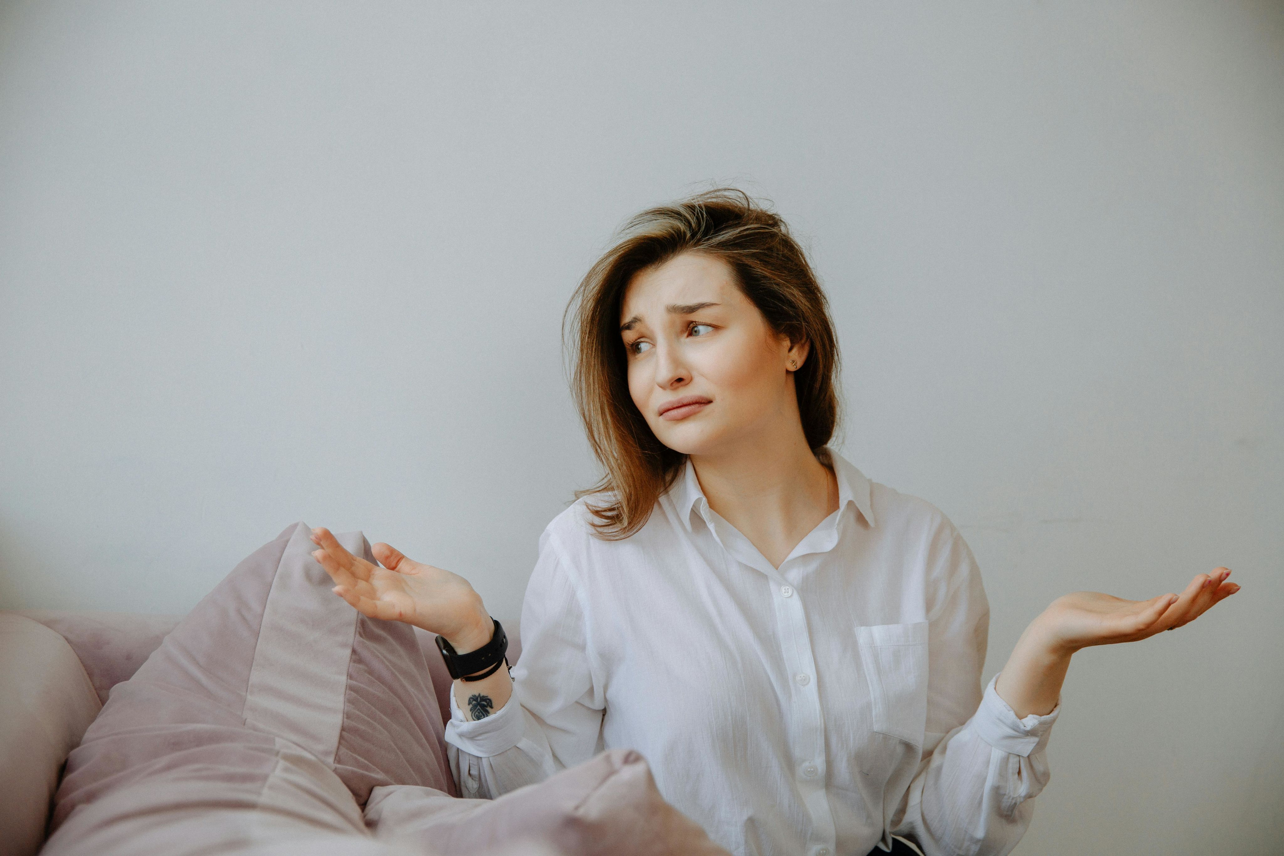 Woman in white shirt sitting indoors on a couch, gesturing with a confused expression, hands up in a shrug.